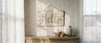 Rustic wooden Hello sign hanging on white brick wall above vintage console table with dried flowers and ceramic pitcher in sunlit room.