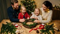 Happy Caucasian family with two young daughters making Christmas wreath together at home with decorated tree and festive lights in background.