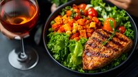 Grilled chicken breast with roasted butternut squash cubes on fresh green lettuce salad, served with red wine in elegant glass on dark background.