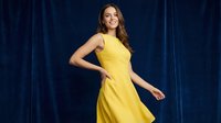 Young Caucasian woman in bright yellow sleeveless dress posing confidently against dark blue velvet curtain background for fashion and lifestyle content.