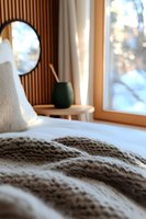 Cozy bedroom interior with knitted blanket, white bedding, round mirror, and green vase on side table near window creating peaceful morning atmosphere.