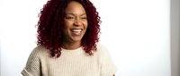 Young African American woman with curly burgundy hair smiling warmly in cream sweater against white background for lifestyle and portrait photography.