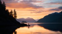 Silhouette of person standing on lake shore during dramatic sunset, with mountains and evergreen trees reflecting in calm water. Peaceful wilderness scene.