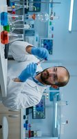 Male scientist with dark hair and beard, South Asian ethnicity, wearing white lab coat and blue nitrile gloves, examining blood sample in test tube in modern laboratory setting with equipment and colorful reagent bottles.