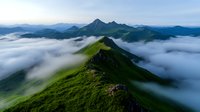 Dramatic mountain ridge rises above low-lying clouds, covered in vibrant green grass. Misty fog flows through valleys creating ethereal atmosphere in mountain landscape.
