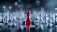 Red glass bishop chess piece stands out among silver pieces on reflective surface with blurred bokeh background creating dramatic leadership concept.