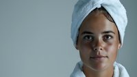 Young mixed race woman wearing white bath towel on head with fresh clean glowing skin after shower, looking at camera with natural expression and minimal makeup.