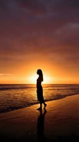 Silhouetted woman standing on beach at sunset, gazing at golden horizon over ocean waves. Contemplative coastal scene for travel, wellness, and lifestyle projects.
