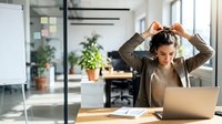 Young professional woman adjusting headphones while working on laptop in modern office workspace with plants and natural light streaming through windows.