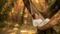 Woman relaxing in hammock strung between trees in forest during golden hour sunlight, peaceful outdoor leisure and nature retreat setting.