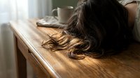 Young woman with long brown hair resting head on wooden table in bright room. Concept of fatigue, stress, exhaustion, mental health, burnout, depression.