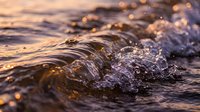 Ocean waves crashing on shore during golden hour sunset with sparkling water droplets and warm light reflections creating peaceful seascape atmosphere.