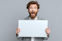 Excited man with curly hair and red beard in gray suit holding blank white poster with surprised expression against neutral background.