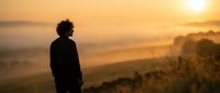 Young African American man silhouette standing on hill overlooking misty valley during golden sunrise, contemplating peaceful morning landscape scenery.