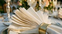 Folded cream napkin with gold napkin ring on formal dining table with blurred glassware and floral centerpiece in background, suitable for event planning and hospitality design.