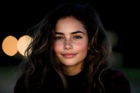 Young Hispanic woman with long dark wavy hair smiling warmly at camera against blurred bokeh background. Natural makeup and casual black top create authentic portrait.
