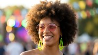 Happy young African American woman with afro hairstyle and sunglasses smiling at colorful festival or carnival with bokeh lights background.