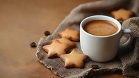 Hot chocolate drink in white ceramic cup with star shaped cookies on rustic burlap cloth, scattered coffee beans creating cozy winter atmosphere.