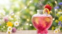 Heart-shaped glass bottle with pink liquid and strawberry garnish on wooden surface with wildflowers and bokeh lights background for romantic occasions.