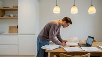 Young Caucasian man working from home at kitchen counter with laptop and documents in modern minimalist interior with pendant lighting.