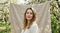 Young Caucasian woman with long brown hair in white clothing posing outdoors against natural linen backdrop with blooming trees in spring garden setting.