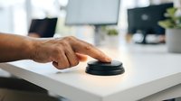 Senior male hand pressing black service bell on white desk in modern office environment for customer assistance and support.