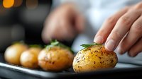 Young potatoes seasoned with herbs and spices on dark background, close up view of hand garnishing roasted baby potatoes with fresh rosemary.