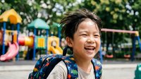 Happy Asian elementary school boy with backpack smiling joyfully at outdoor playground during sunny day, expressing childhood excitement and carefree moments.