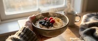 Hands holding bowl of oatmeal porridge topped with fresh berries and nuts by sunny window, warm breakfast scene for food and wellness content.