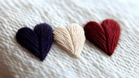 Colorful embroidered hearts in purple, beige, and red thread on white textured fabric, showcasing handcrafted needlework detail.