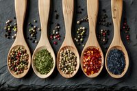 Variety of colorful spices and seasonings in wooden spoons arranged on black slate background, including peppercorns, dried herbs, chili flakes and poppy seeds for cooking and food styling.