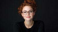 Young redhead woman with glasses and updo hairstyle wearing black top smiling confidently against dark background. Professional headshot portrait with natural expression.