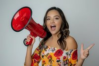 Young Asian woman in floral off-shoulder dress holds red megaphone while showing peace sign against light background, expressing excitement and positive energy.