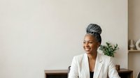 Mature woman with gray hair wearing white blazer and black top, smiling while looking away in modern office setting with plant decor.