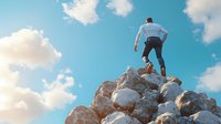 Young Caucasian businessman in formal attire stands triumphantly on mountain peak against blue sky with clouds, representing achievement and leadership success.