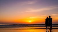 Romantic couple silhouettes standing on beach watching golden sunset over ocean waves with colorful sky reflecting on wet sand creating peaceful moment.