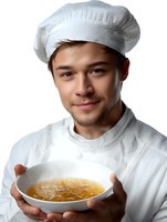 Young male chef in white uniform and toque holding bowl of fresh homemade soup against white background.