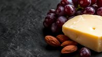 Fresh parmesan cheese wedge with red grapes and raw almonds arranged on dark stone background, selective focus on cheese with copy space.