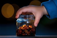 Hand protecting glass jar filled with golden coins against dark background with warm bokeh lights creating magical atmosphere for savings concept.