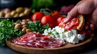 Mediterranean antipasto platter with cured meats, feta cheese, fresh herbs, olives and tomatoes on rustic wooden board, selective focus on foreground.