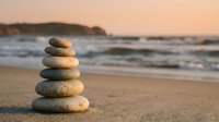 Balanced stone cairn on sandy beach at sunset with ocean waves and coastal cliffs creating peaceful zen meditation atmosphere for wellness and mindfulness concepts.
