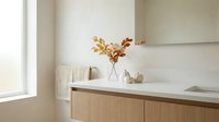 Modern minimalist bathroom vanity with wooden cabinets, white countertop, decorative autumn branches in glass vase, neutral beige walls, natural lighting.