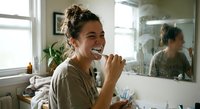 Young mixed-race woman brushing teeth in modern bathroom mirror reflection morning routine dental hygiene self-care wellness lifestyle healthy habits.