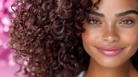 Young mixed-race woman with curly brown hair smiling at camera against pink floral background for beauty and lifestyle content.