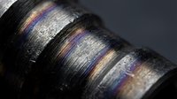 Close-up macro shot of metallic surface with iridescent rainbow oil slick patterns creating colorful interference bands on dark industrial material.