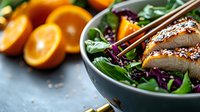 Grilled sesame chicken breast on fresh green salad with red cabbage and orange slices in dark ceramic bowl, healthy lunch concept on stone background.