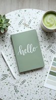 Sage green notebook with hello lettering on terrazzo desk, matcha latte, succulent, paper clips, and color swatches in flat lay style.