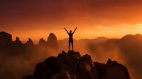 Silhouette of triumphant hiker with raised arms celebrating success on mountain peak at golden sunset with dramatic orange sky and layered mountain ranges.