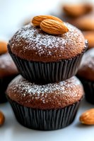 Chocolate cupcakes topped with almond and powdered sugar in black paper cups, stacked on white surface with scattered nuts, macro food photography.