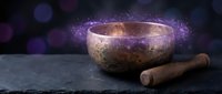 Tibetan singing bowl with wooden mallet on dark slate surface surrounded by mystical purple bokeh lights for meditation and sound healing practices.